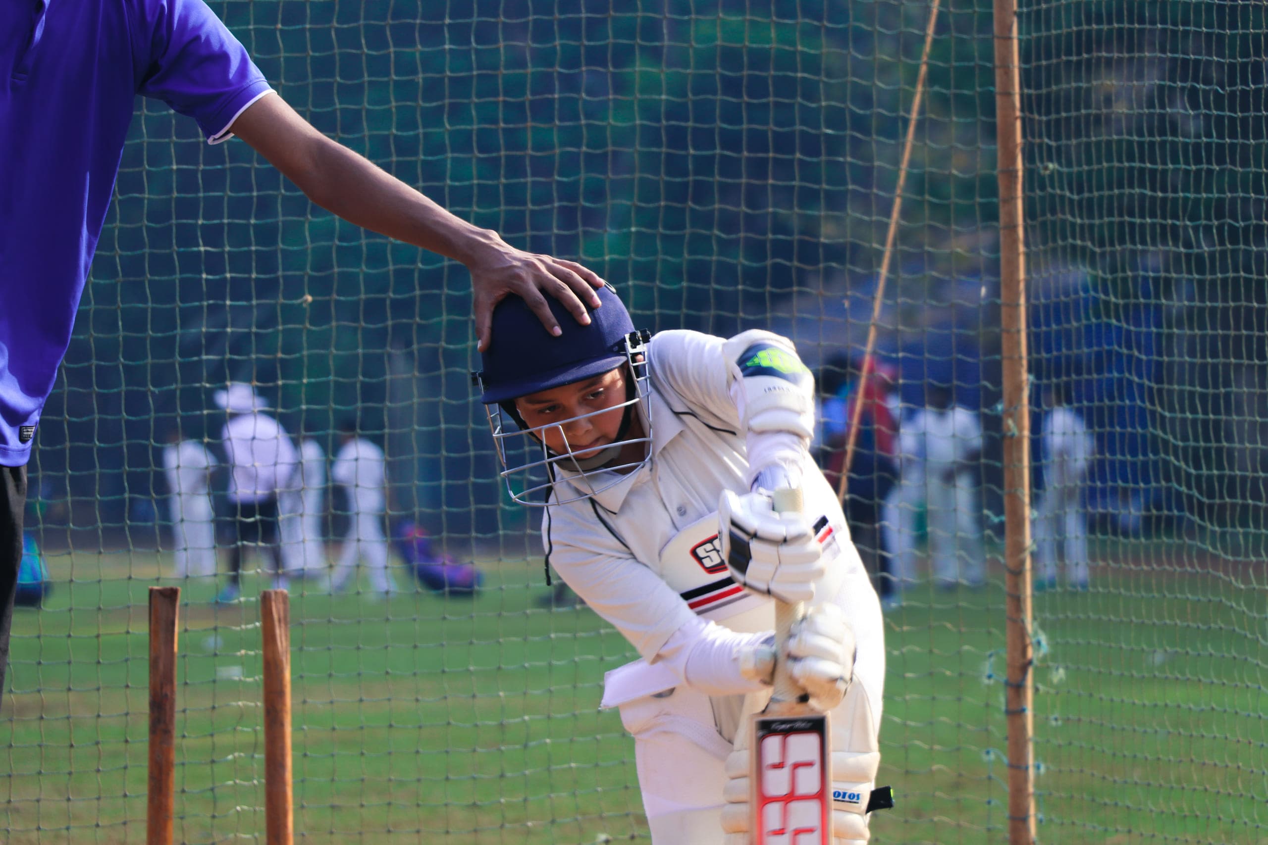A young batsman defending in the nets while a coach steadies his helmet